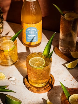 Glass of Tepache with a lime slice and pineapple leaf garnish, surrounded by Casa Piñata's Tepache bottle and fresh lime wedges on a sunlit surface